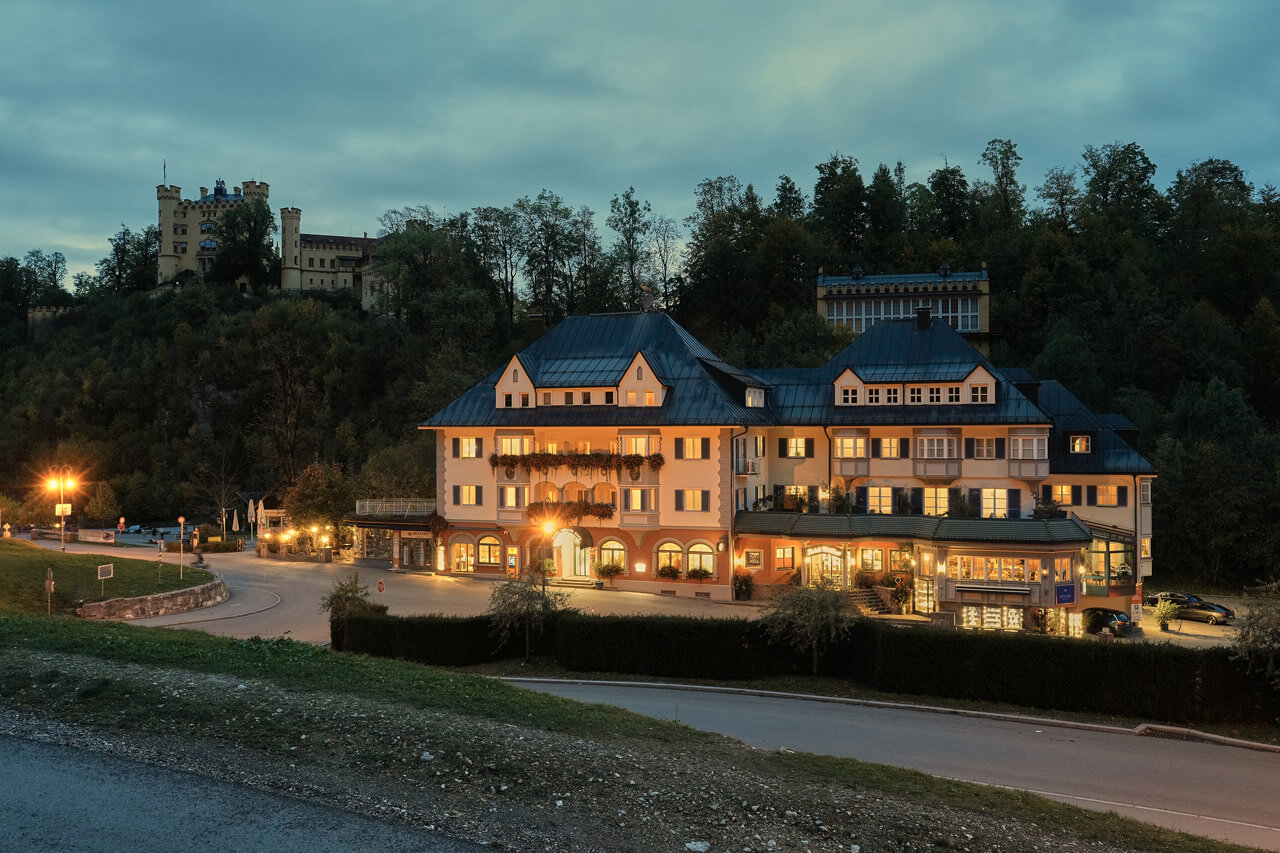 Beleuchtete Hotel Müller mit dunklem Dach steht in der Dämmerung neben einem bewaldeten Hügel. Im Hintergrund ist auf der Hügelkuppe Hohenschwangauzu erkennen.