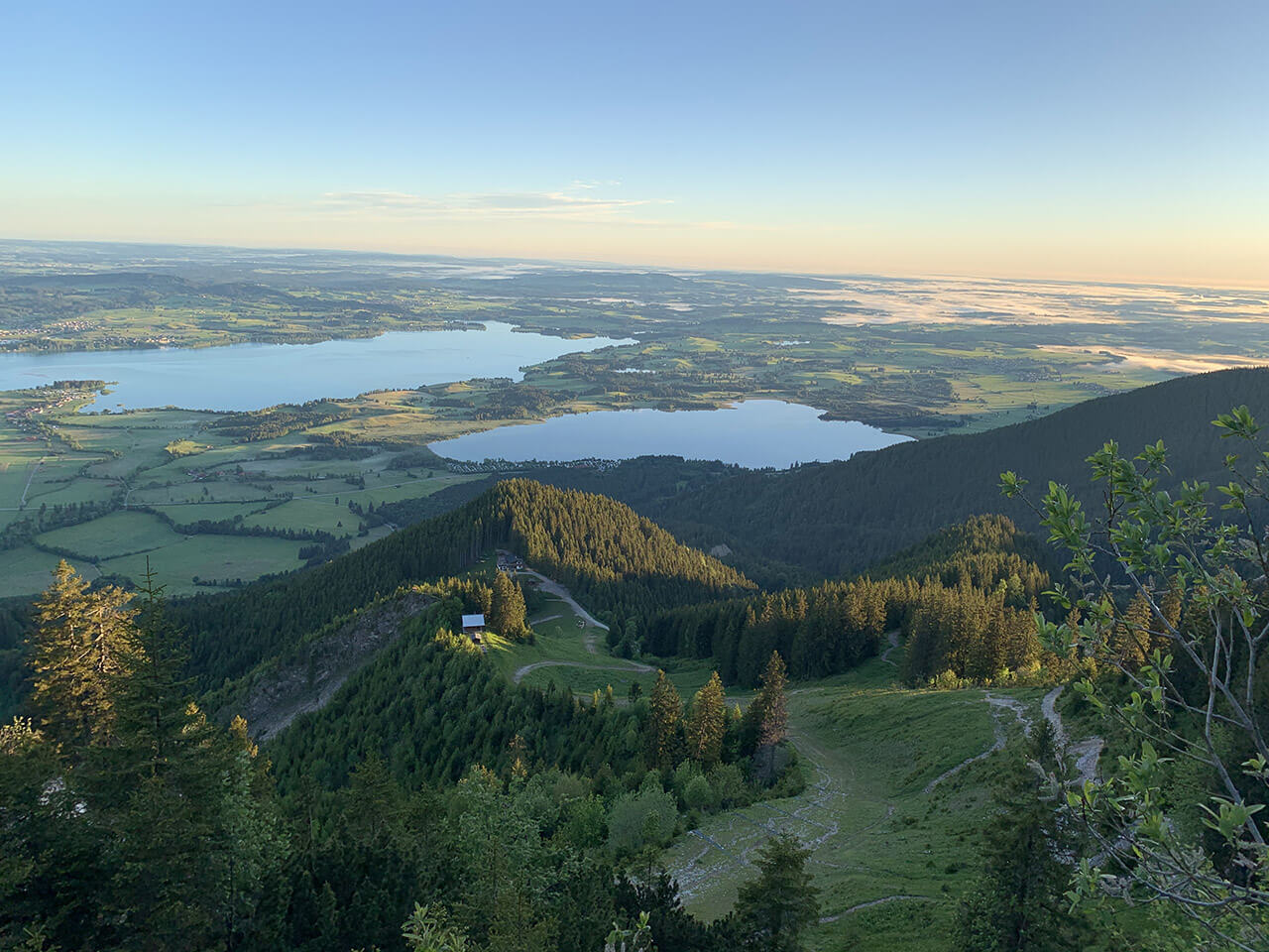 Eine malerische Aussicht auf ein bewaldetes Tal mit einem kleinen See in der Ferne, umgeben von hoch aufragenden Bergen unter einem klaren blauen Himmel.