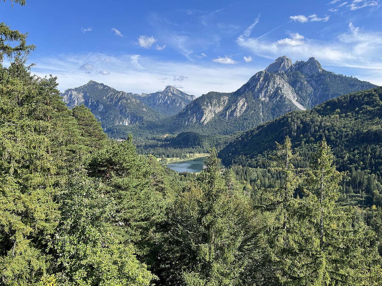 Eine malerische Aussicht auf ein bewaldetes Tal mit einem kleinen See in der Ferne und dem Kalvarienberg im Hintergrund unter einem klaren blauen Himmel.