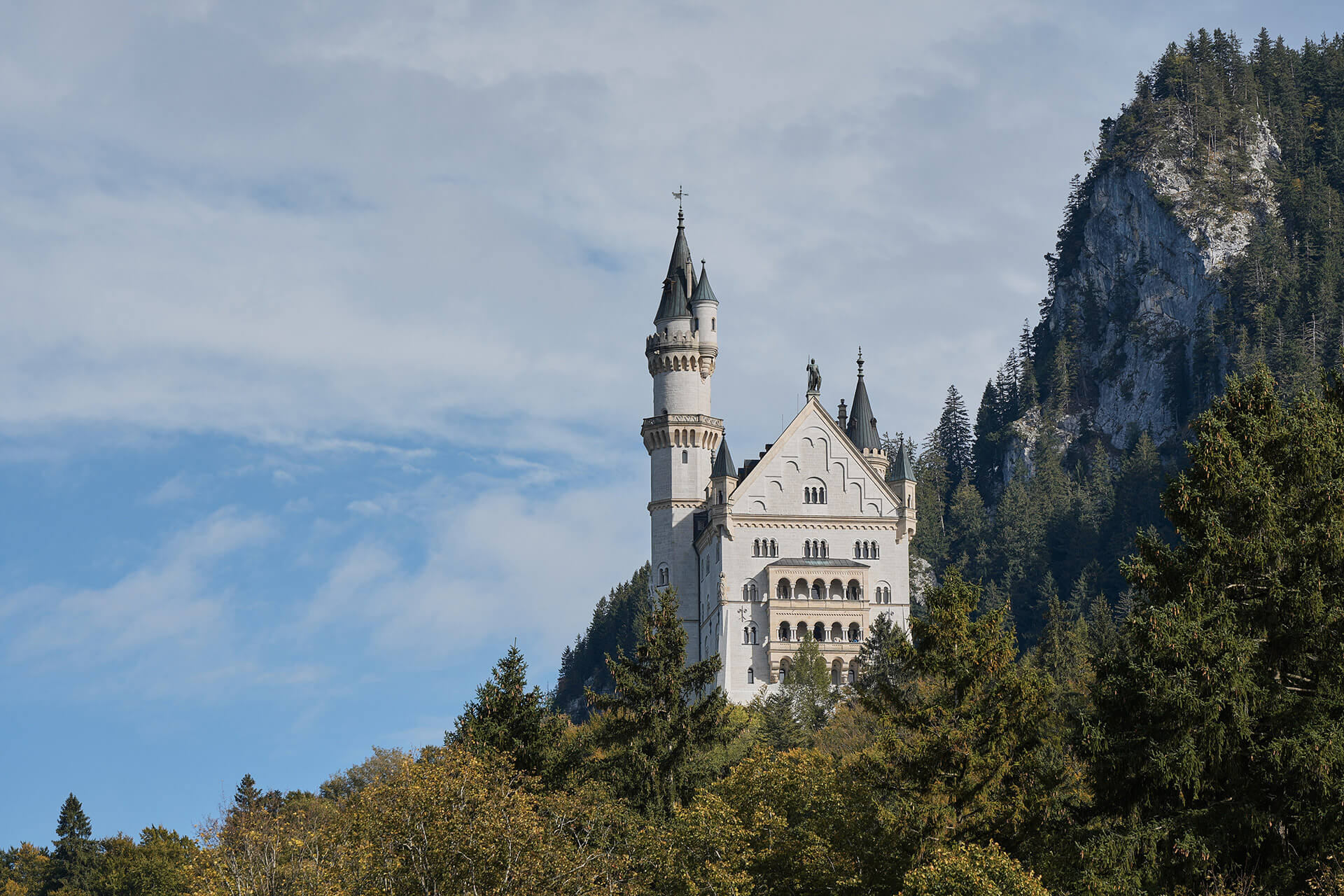 Das Schloss Neuschwanstein mit hohen Türmen und kunstvollen Details liegt vor der Kulisse bewaldeter Hügel und eines teilweise bewölkten Himmels.